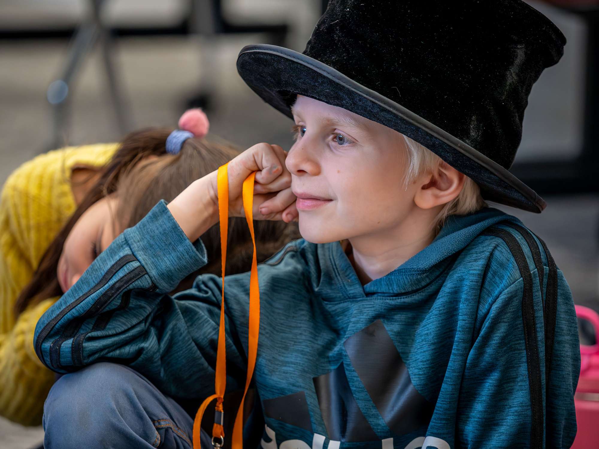 "A child wearing a large black top hat and teal hoodie sits on the floor holding an orange lanyard, while another child in a yellow sweater leans nearby."