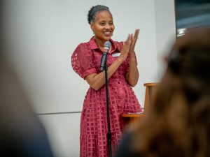 Vice President for Development and Alumni Engagement Rita Walters, wearing a maroon dress, stands in front of a microphone, she is clapping.