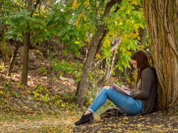 Photo of a student leaning against a tree and writing in a notebook
