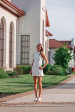 A young woman in a white dress standing proudly on a college campus with a white building in the background.