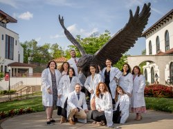 Eleven students wearing white lab coats pose for a photo in front of a bronze red hawk statue.