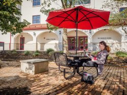 A student sitting outside of the School of Nursing and Graduate School reading a book on a sunny day under a table umbrella.