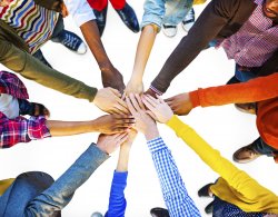 Photo of about 10 individuals in a circle with their hands together symbolizing teamwork.