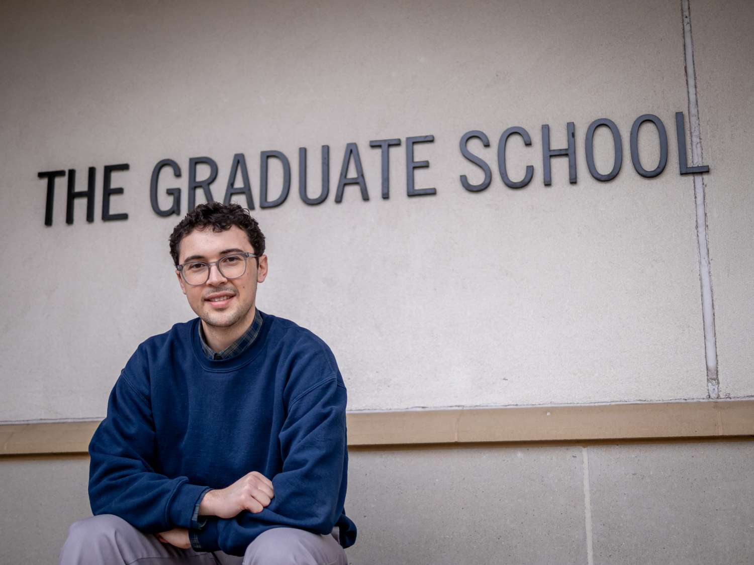 Jake Hutchison sitting in front of a wall with text, The Graduate School.