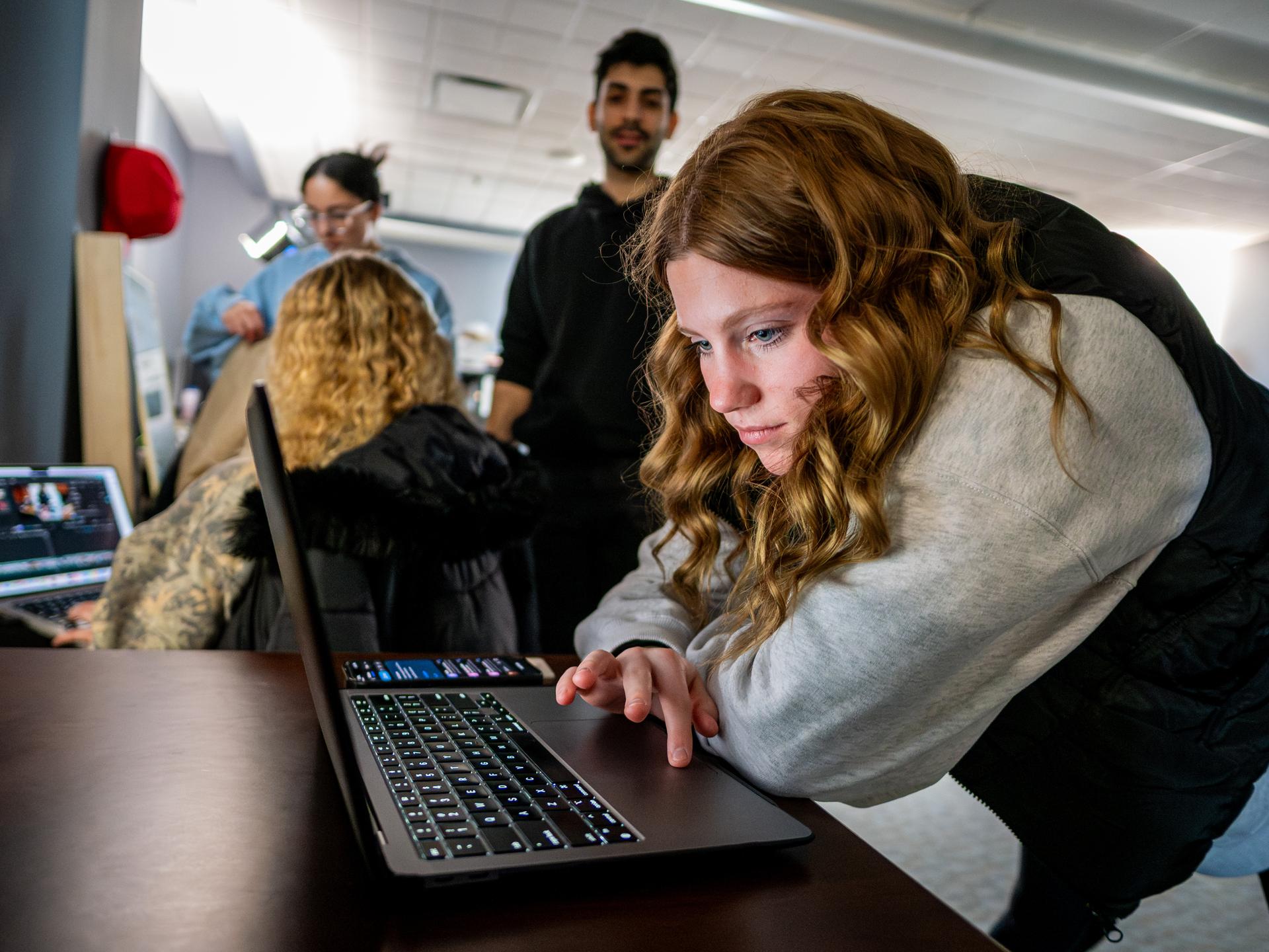 A female student working on a laptop with other students in the background.