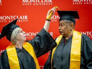 Two women in black gowns, yellow sahes, and black graduation caps. One is fixing the cap of the other.
