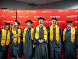 Individuals stand in a line, robed in black gowns with yellow sashes and graduation caps.