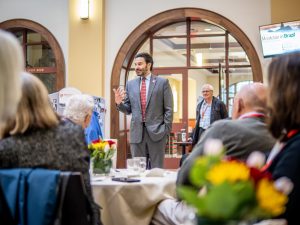 A man in a blue suit and red tie addresses an audience sitting in front of him
