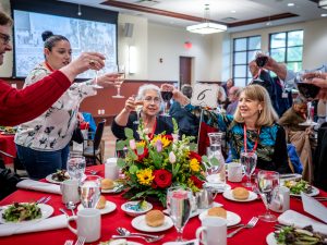 A table of women toast with glasses of wine. The table is covered in a red cloth and has flowers and many plates on it.