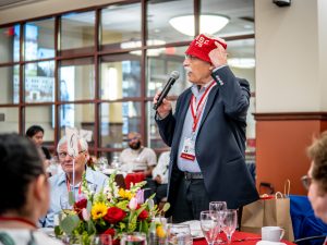A man in a blue suit holds a microphone, addressing a room. He is wearing a red bucket cap with "MSU 1975" written on it.
