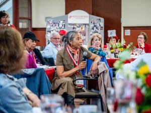 A woman is seated between others at a table; she is holding a microphone to her mouth and is looking to her left.