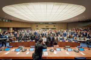 A large crowd gathered and posting for a photo in a United Nations lecture hall. A photographer at work is in the foreground.