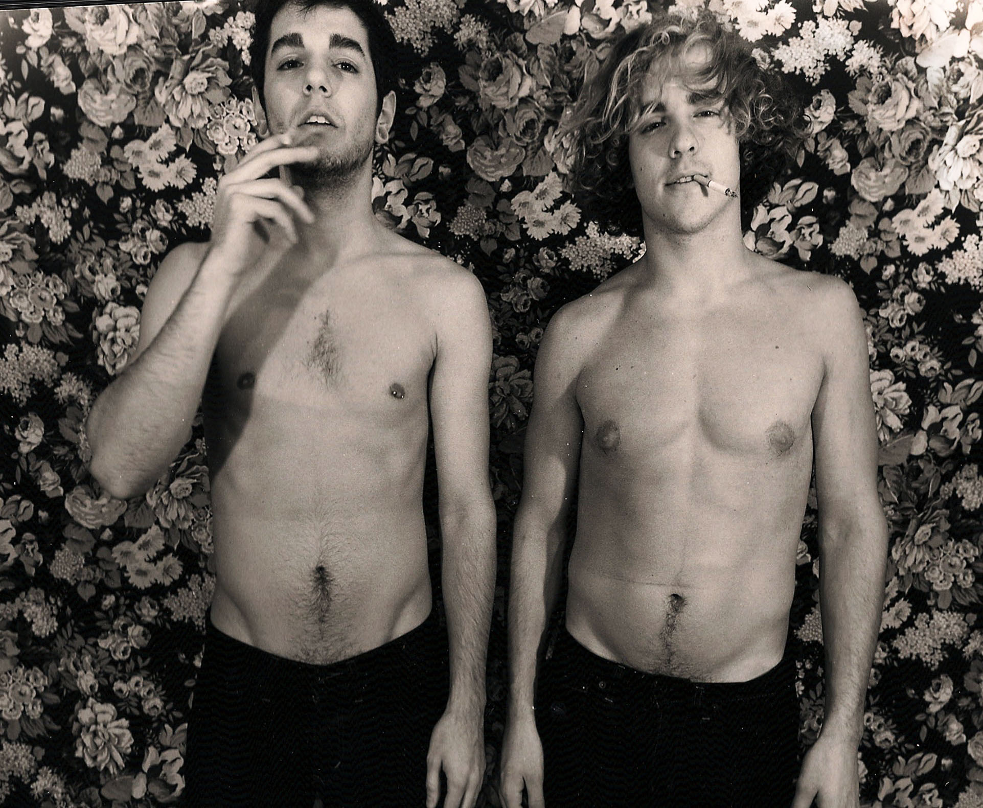 photo of two shirtless men in front of flower pattern wallpaper