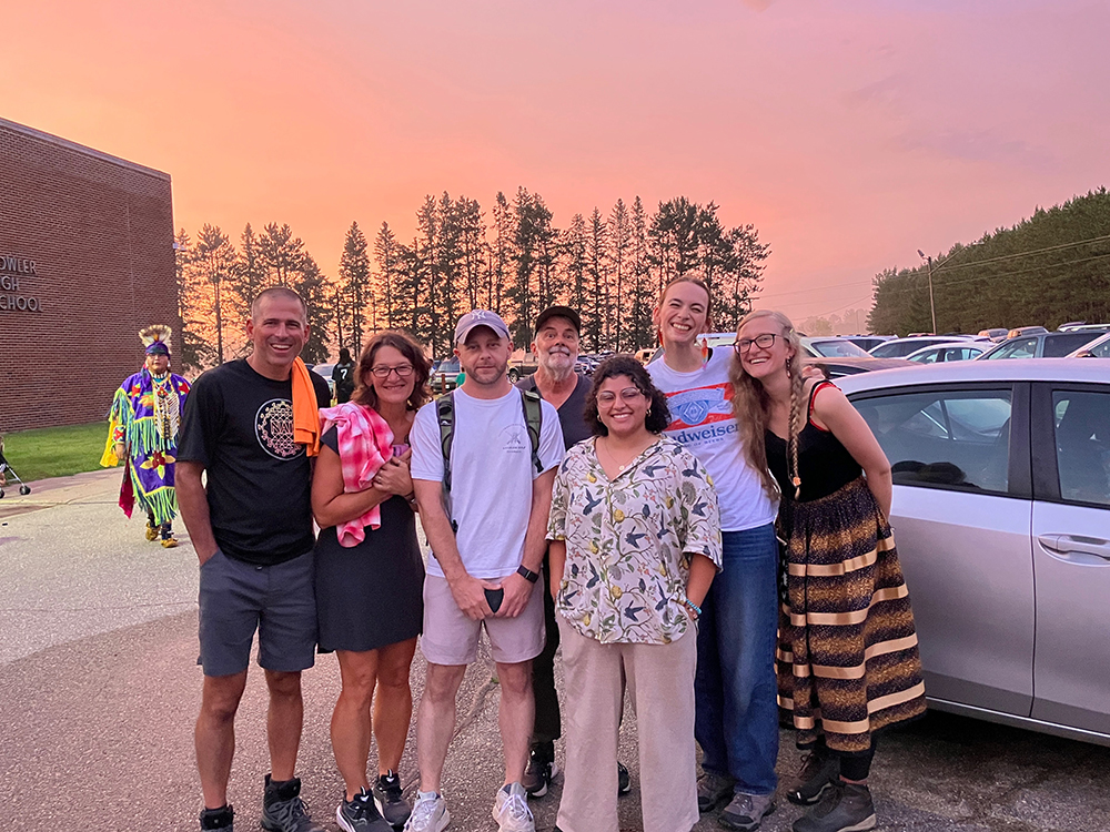 a group stands together smiling with pink sunset behind. A man in traditional Native American celebratory dress can be seen in background