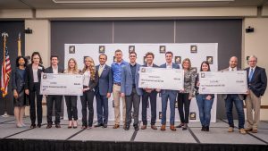 Students stand on stage with oversized checks, and the judges.