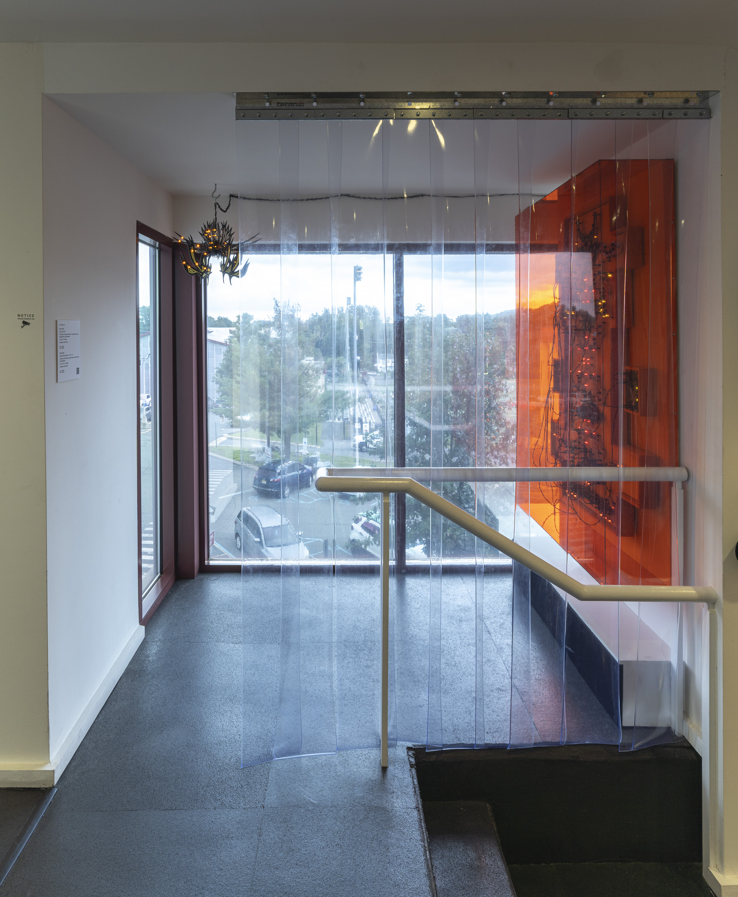 Photo of the Sound Booth installation. On the left is an artwork made of circuit boards and wires behind orange acrylic is affixed to the wall. On the right hangs a small, black lamp. A clear curtain separates the Sound Booth from the rest of its location, which is a parking garage.