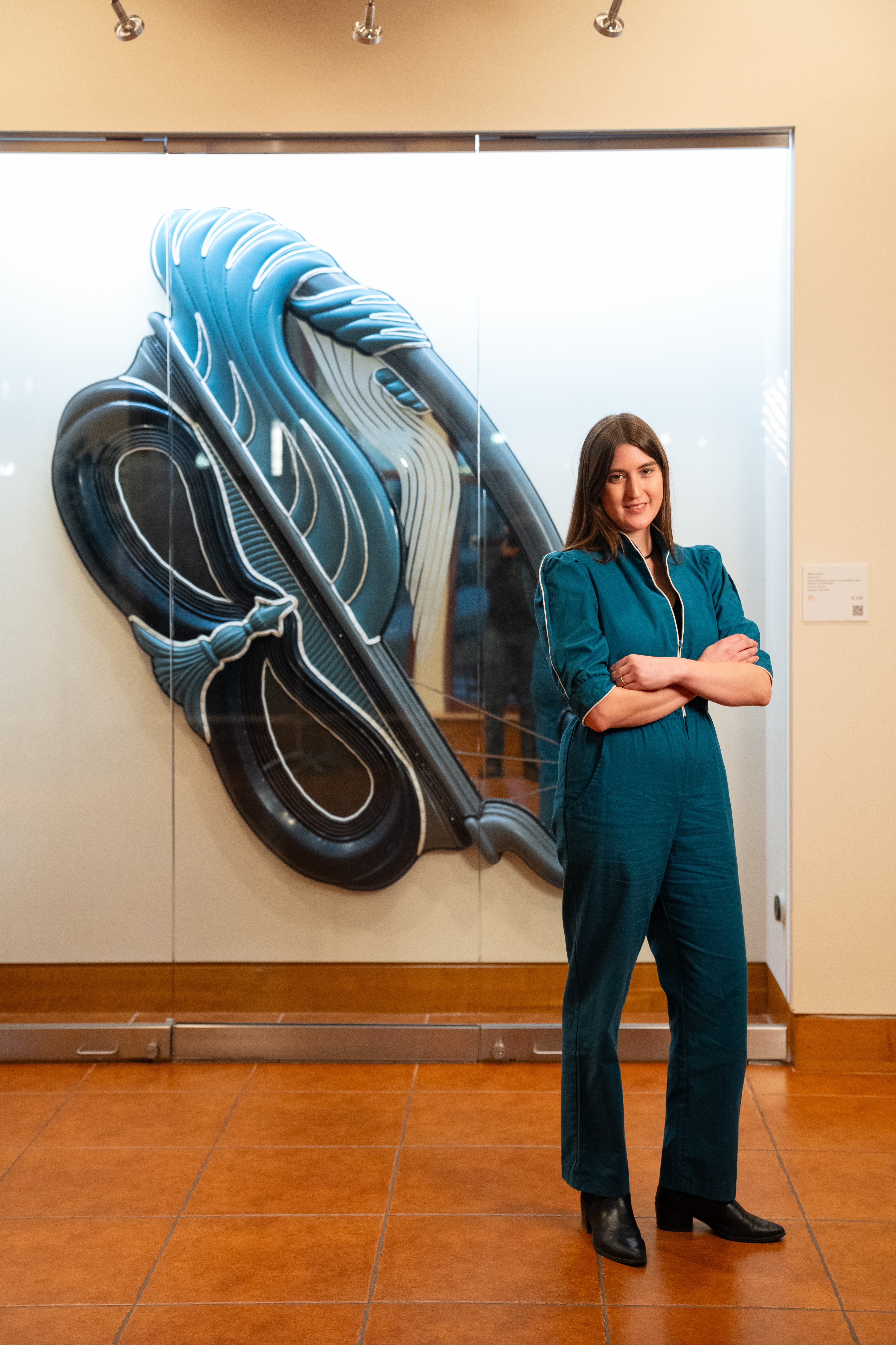 Artist Hilliary Gabryel in a blue jumpsuit stands in front of a blue artwork behind a glass case in the Kasser Theater lobby.