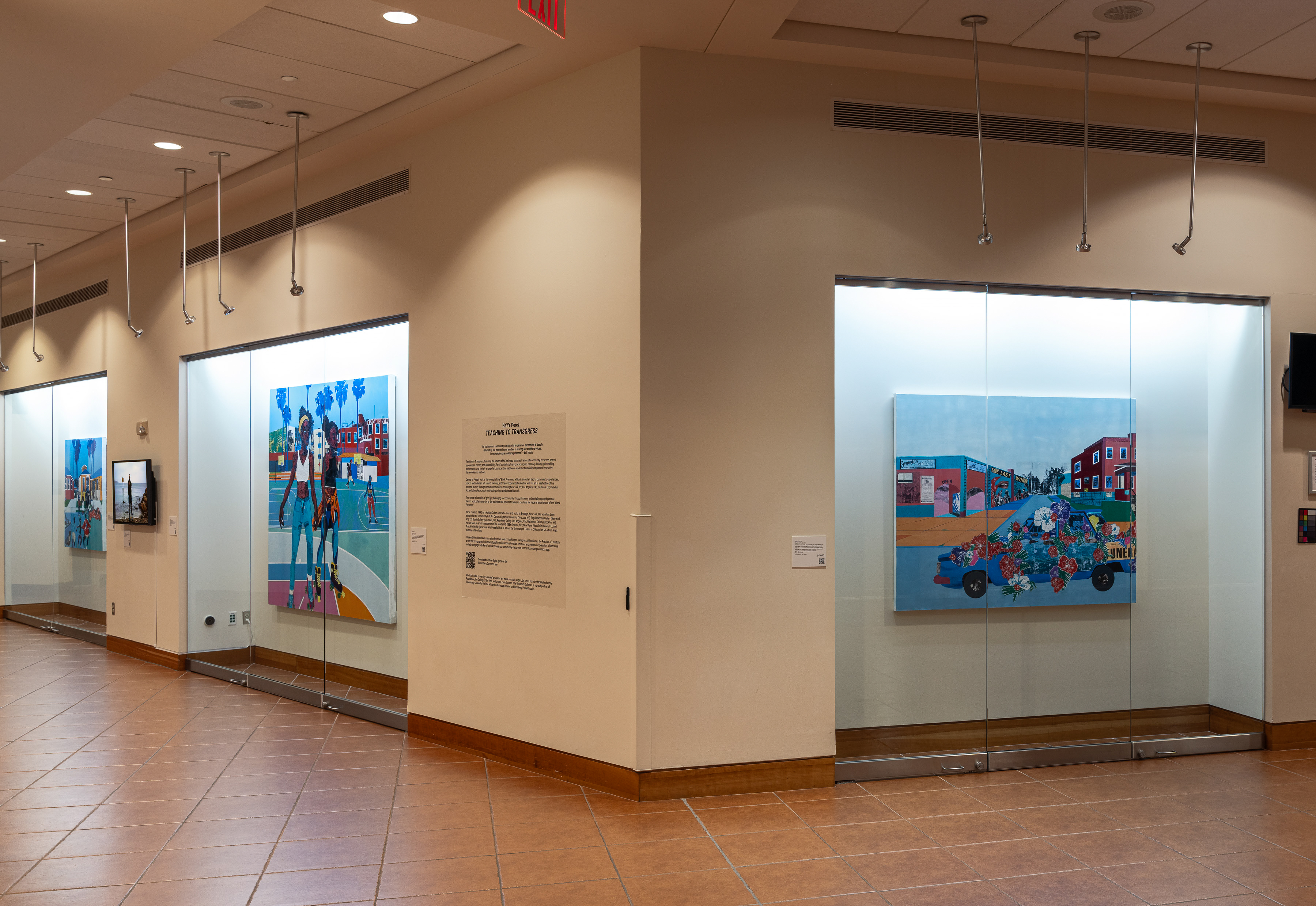 Installation photo of Na'Ye Perez: Teaching to Transgress in the Kasser Theater lobby. The exhibition features large, multicolored paintings in glass display cases.