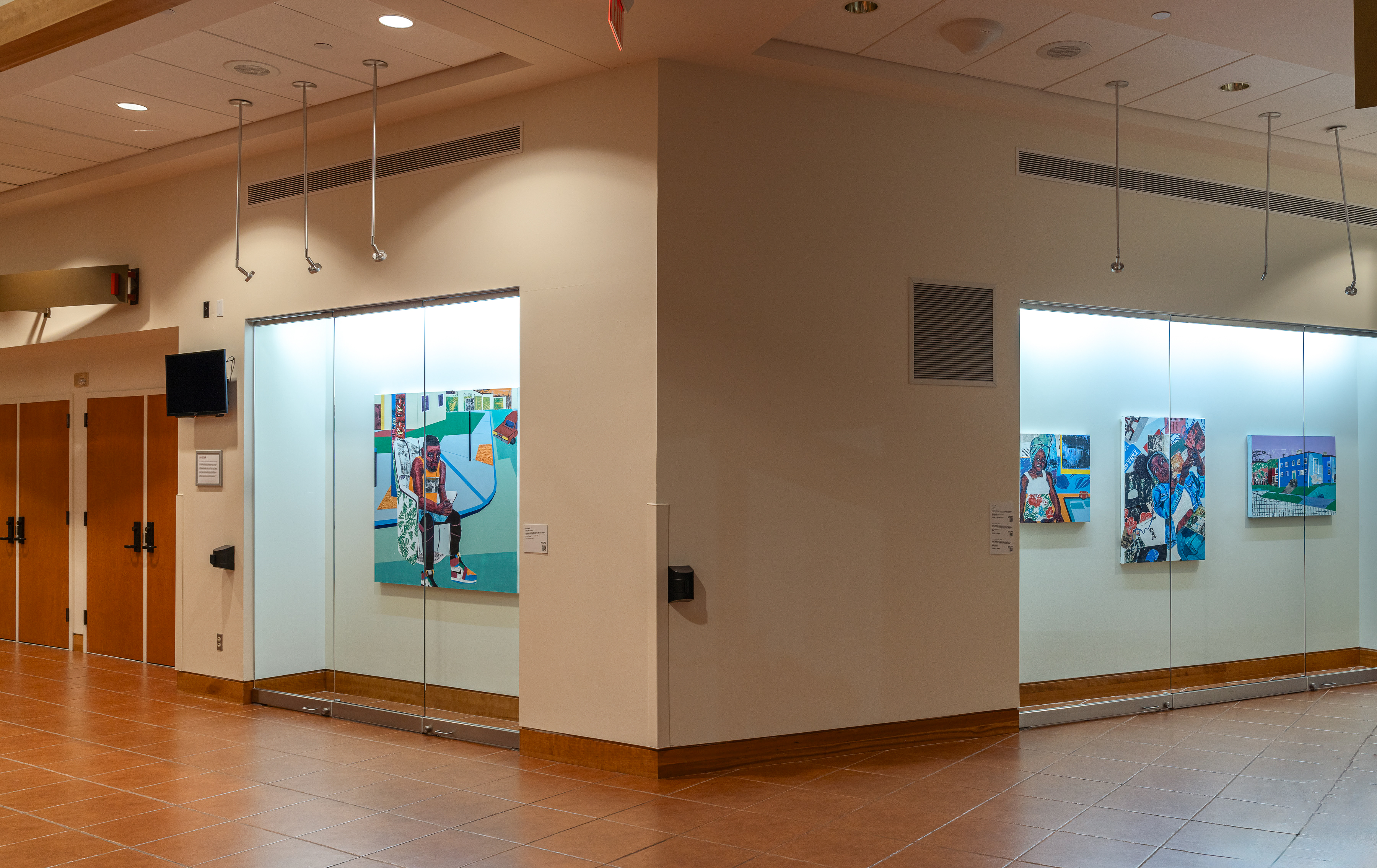 Installation photo of Na'Ye Perez: Teaching to Transgress in the Kasser Theater lobby. The exhibition features large, multicolored paintings in glass display cases.