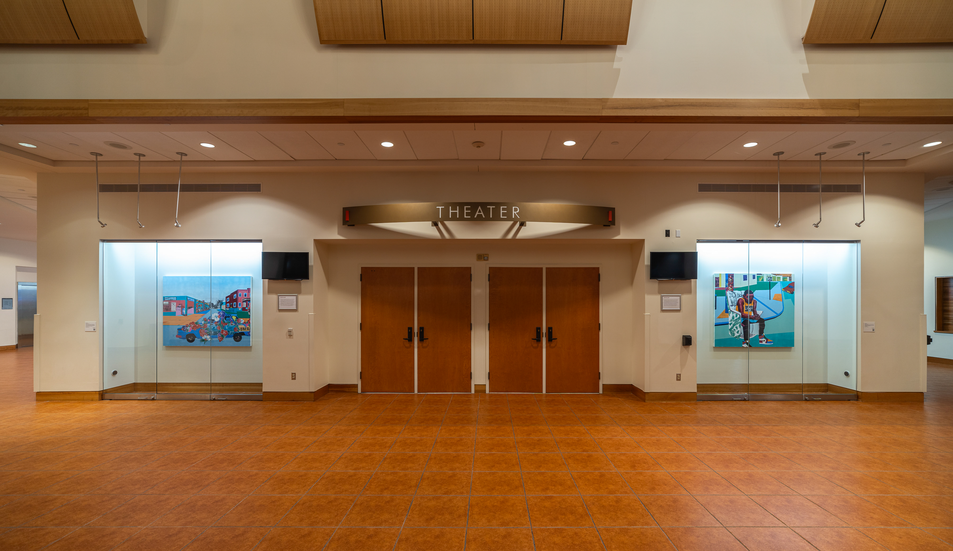 Installation photo of Na'Ye Perez: Teaching to Transgress in the Kasser Theater lobby. The exhibition features large, multicolored paintings in glass display cases.