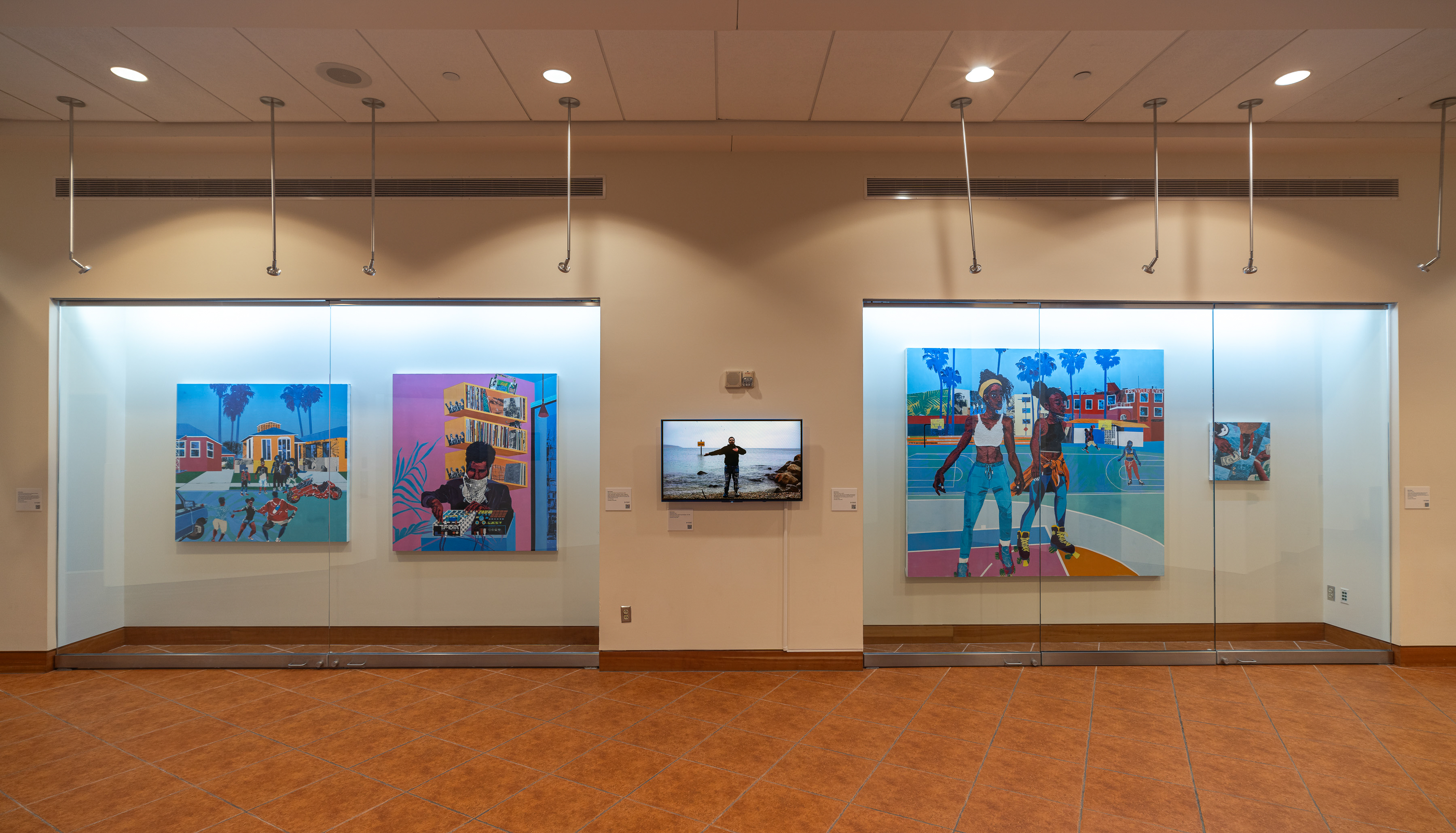 Installation photo of Na'Ye Perez: Teaching to Transgress in the Kasser Theater lobby. The exhibition features large, multicolored paintings in glass display cases.