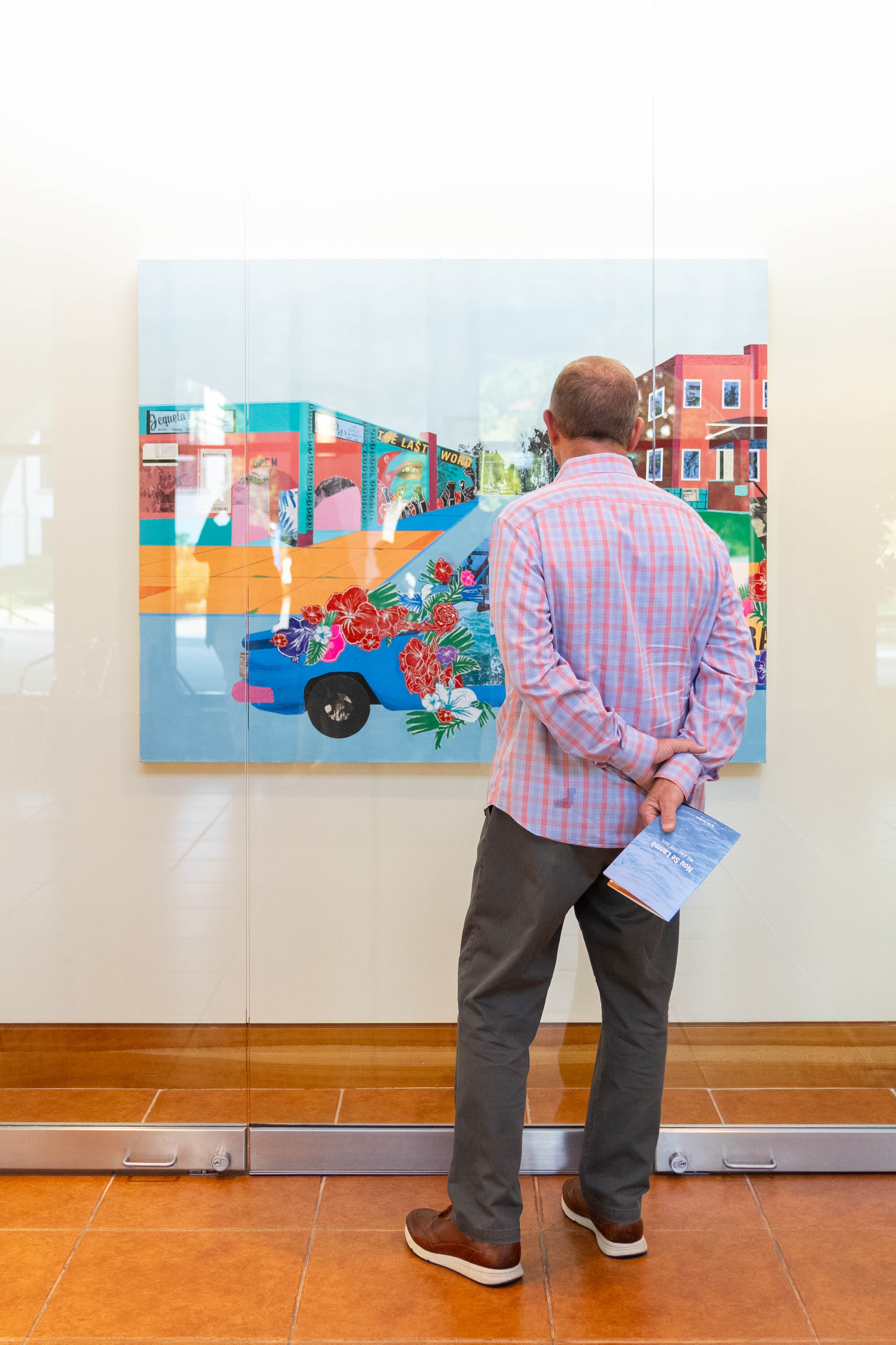 A visitor stands before a multicolored painting behind glass. The visitor's arms are behind their back. The vistior holds a blue booklet.