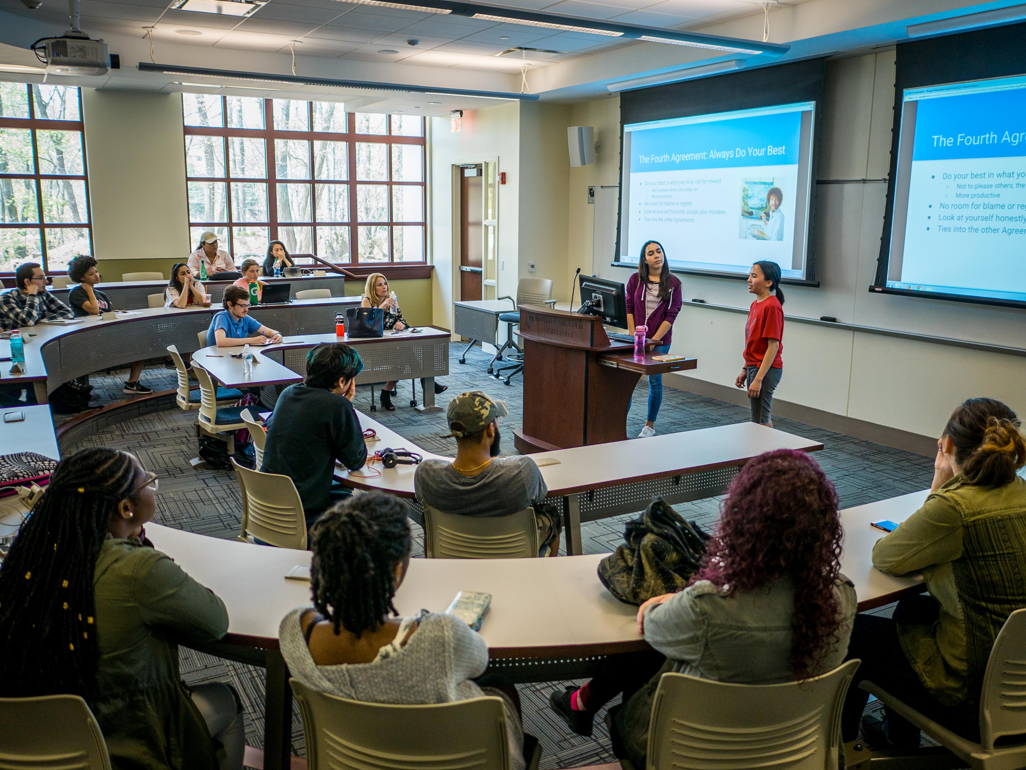 Photo of students presenting in classrrom in front of slides and other students.