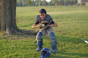Griffin Shoemaker as a young boy, wearing baseball catchers equipment ouside while smiling