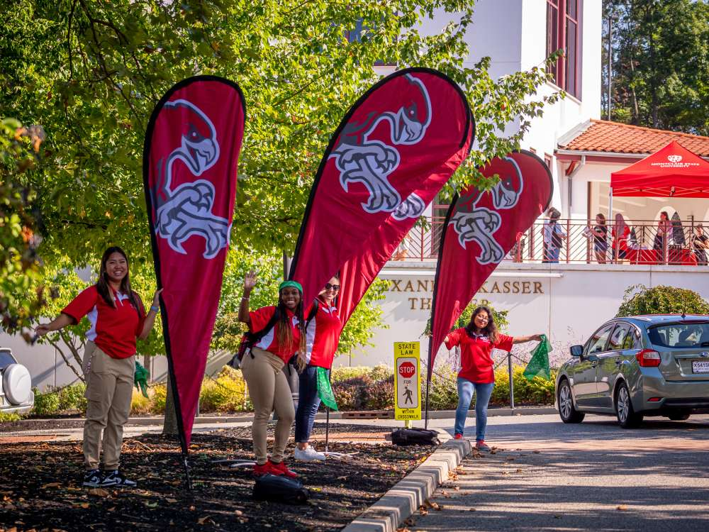 Three Orientation Leaders in red stand beside tall flags while welcoming students to Montclair State University