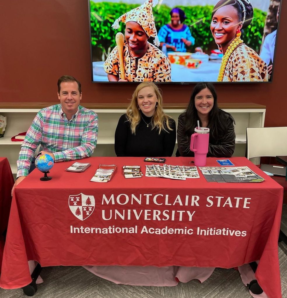 Group of three members of the Office of International Academic Initiatives in front of television with image of Burkina Faso cultural clothing. There is a man on the left side and two women to his right sitting in front of a table with a red tablecloth that has the office logo. 