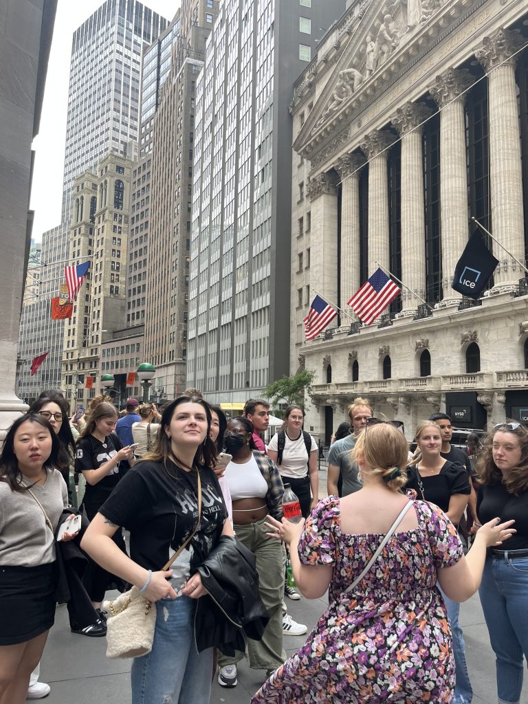 Group of students from different cultures and countries standing with their advisor on Wall Street who explains the history of the area