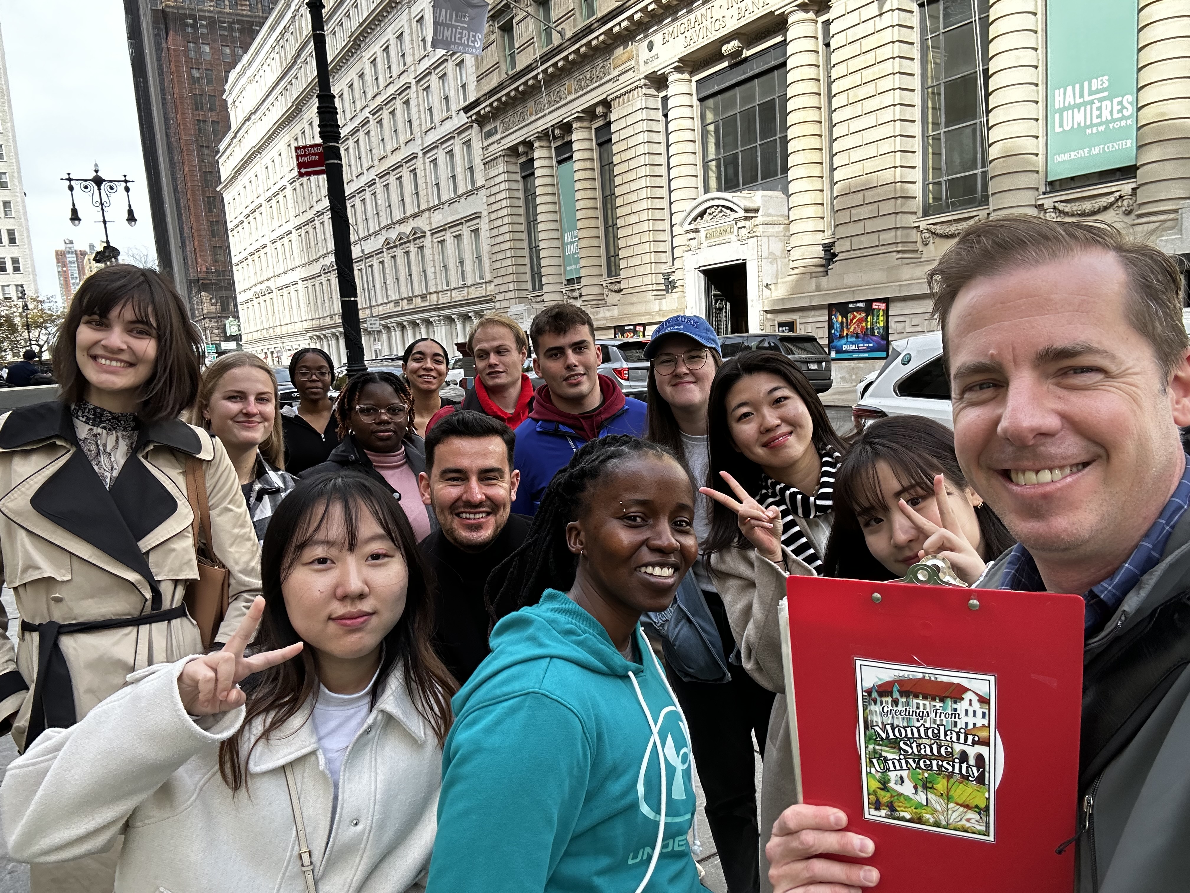 Group of students from different countries and cultures stand together in New York City with a man holding a red clipboard that says "Montclair State University" on the back