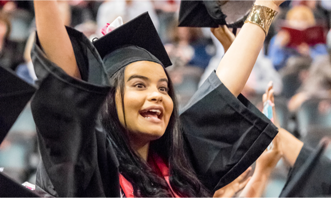 Female Montclair State University student clad in graduation gown raising hands in the air.