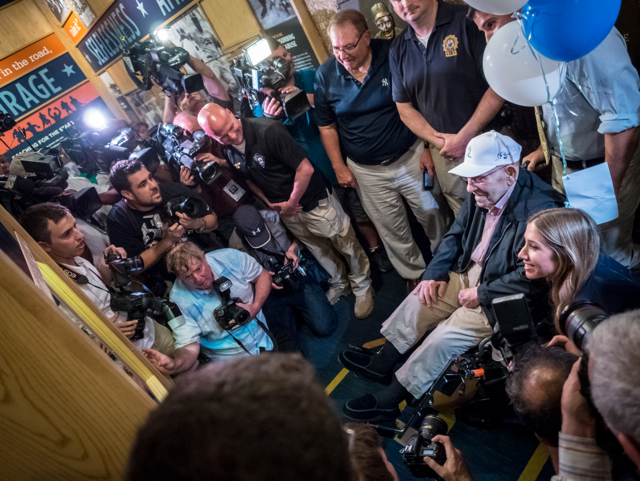 Yogi Berra in wheelchair surrounded by media and students on his 90th birthday.