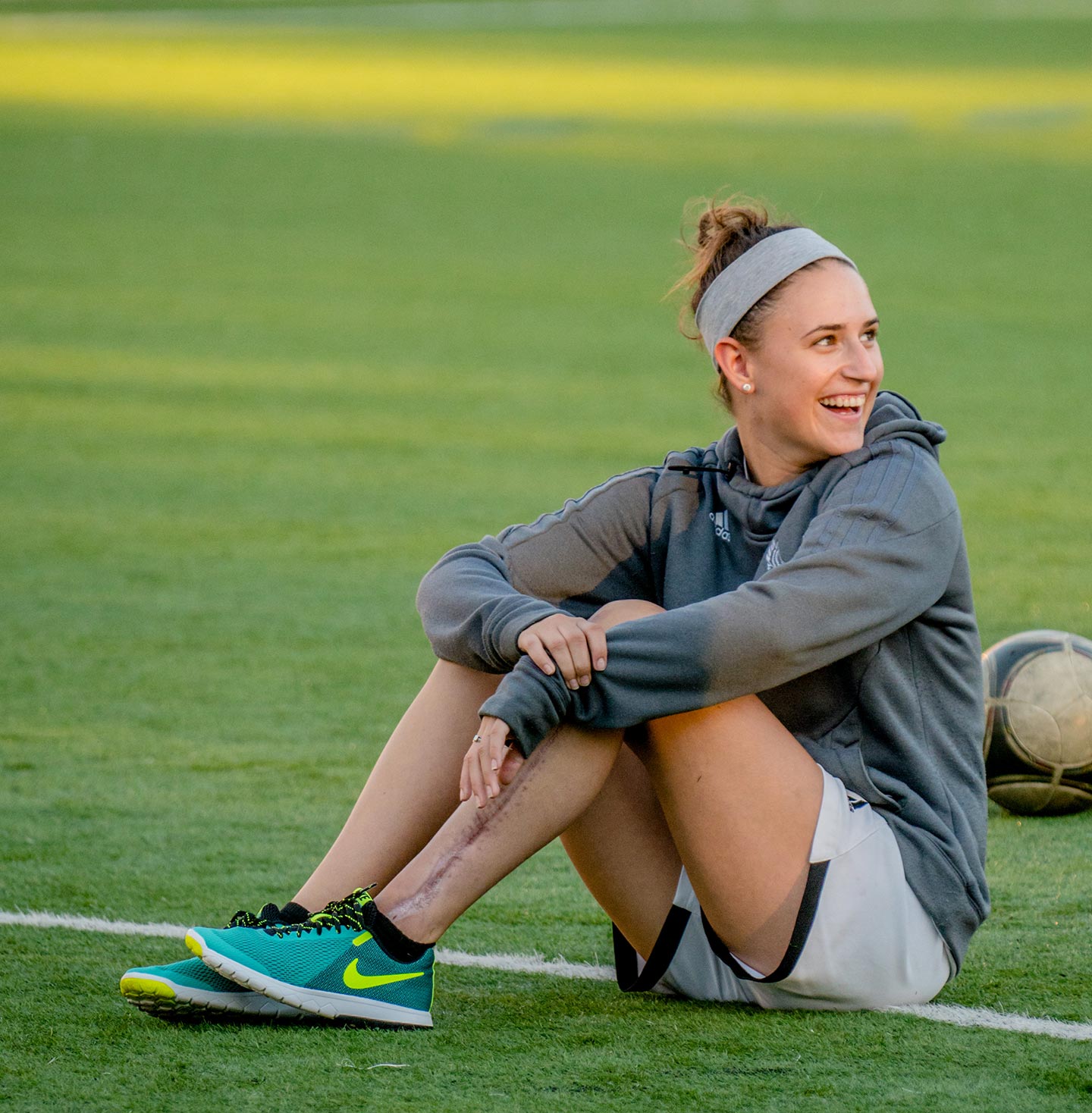 Kim Mignone sitting on soccer field, smiling. Scar on her leg is visible.