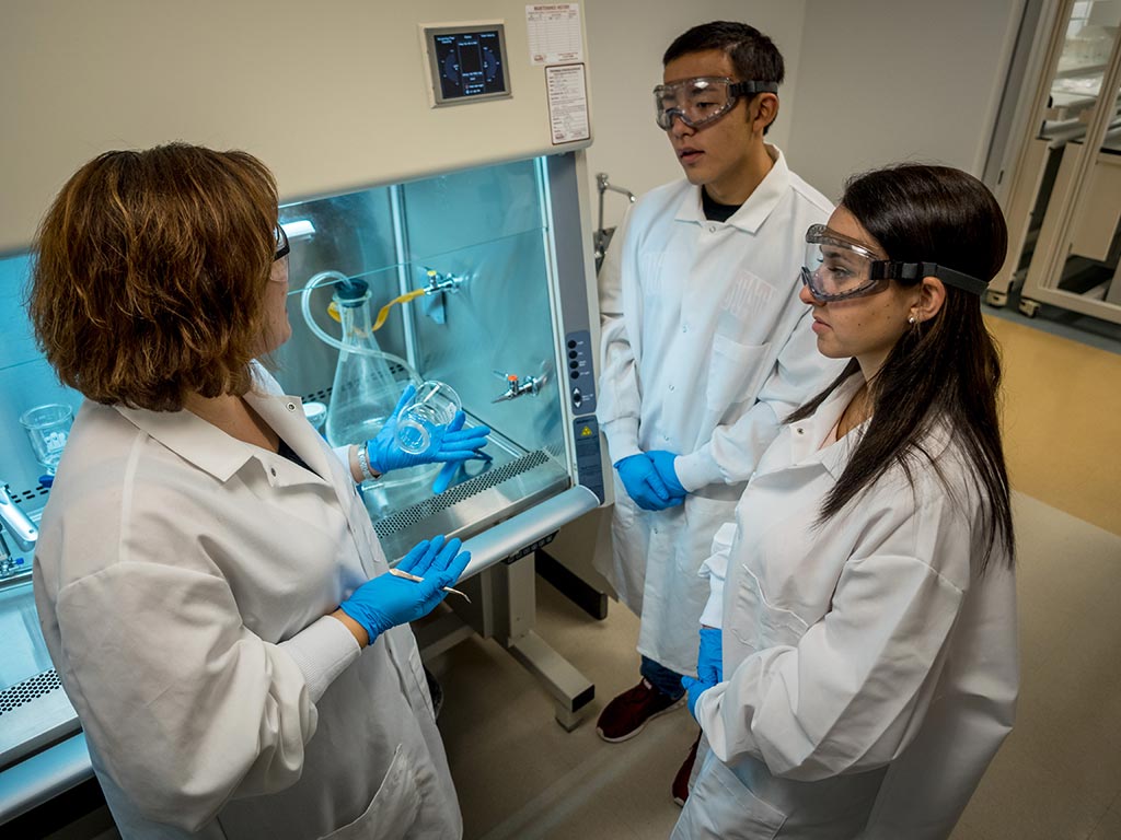 Montclair State University CELS students – wearing safety goggles and lab coats – chatting.