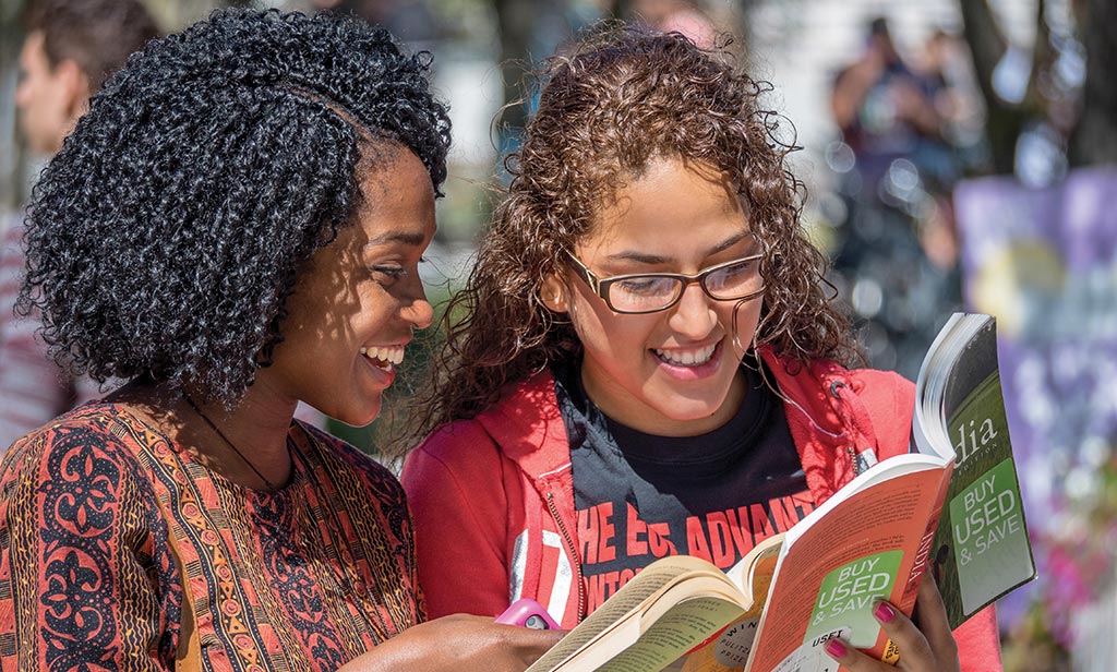 Photo of a pair of female Montclair State University students chatting and reading books together.