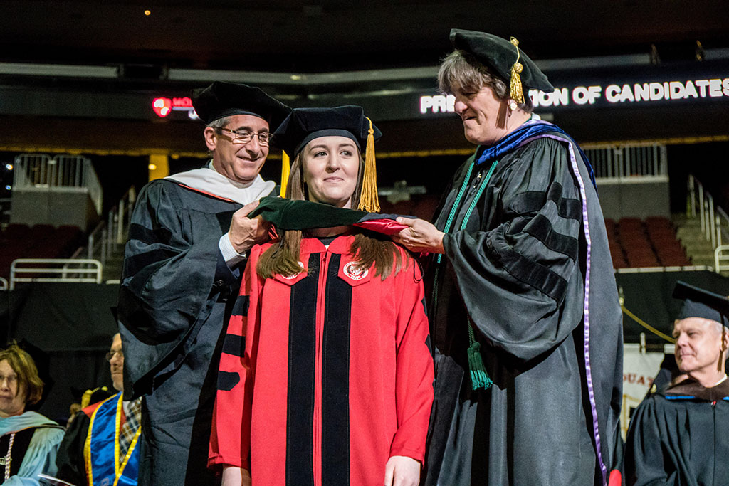 Graduate school student at commencement standing on stage being honored by dean and professor.