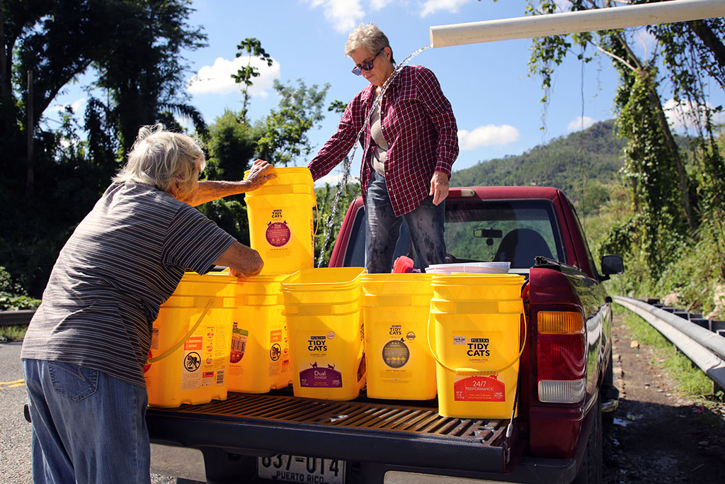 Two women collect water in cat litter buckets from a pipe bringing untreated water from the mountainside outside Utuado, Puerto Rico, three months after Hurricane Maria.
