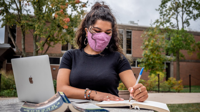 Student wearing mask writing in notebook outside in front of laptop