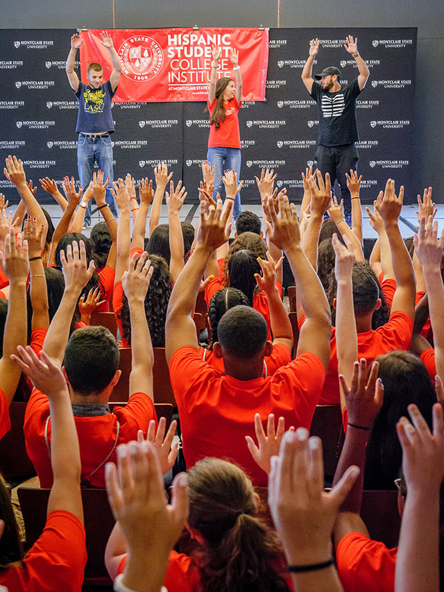 Students of Hispanic Student College Institute raising hands in air with performers on stage doing the same in front of them.