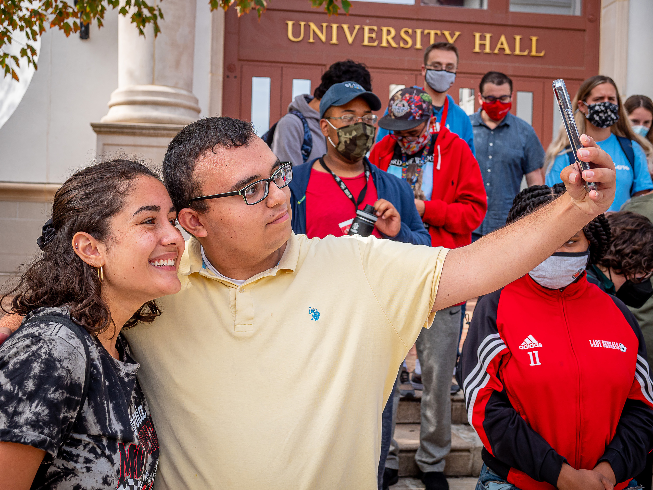 Two students pose for a selfie together with more students in the background