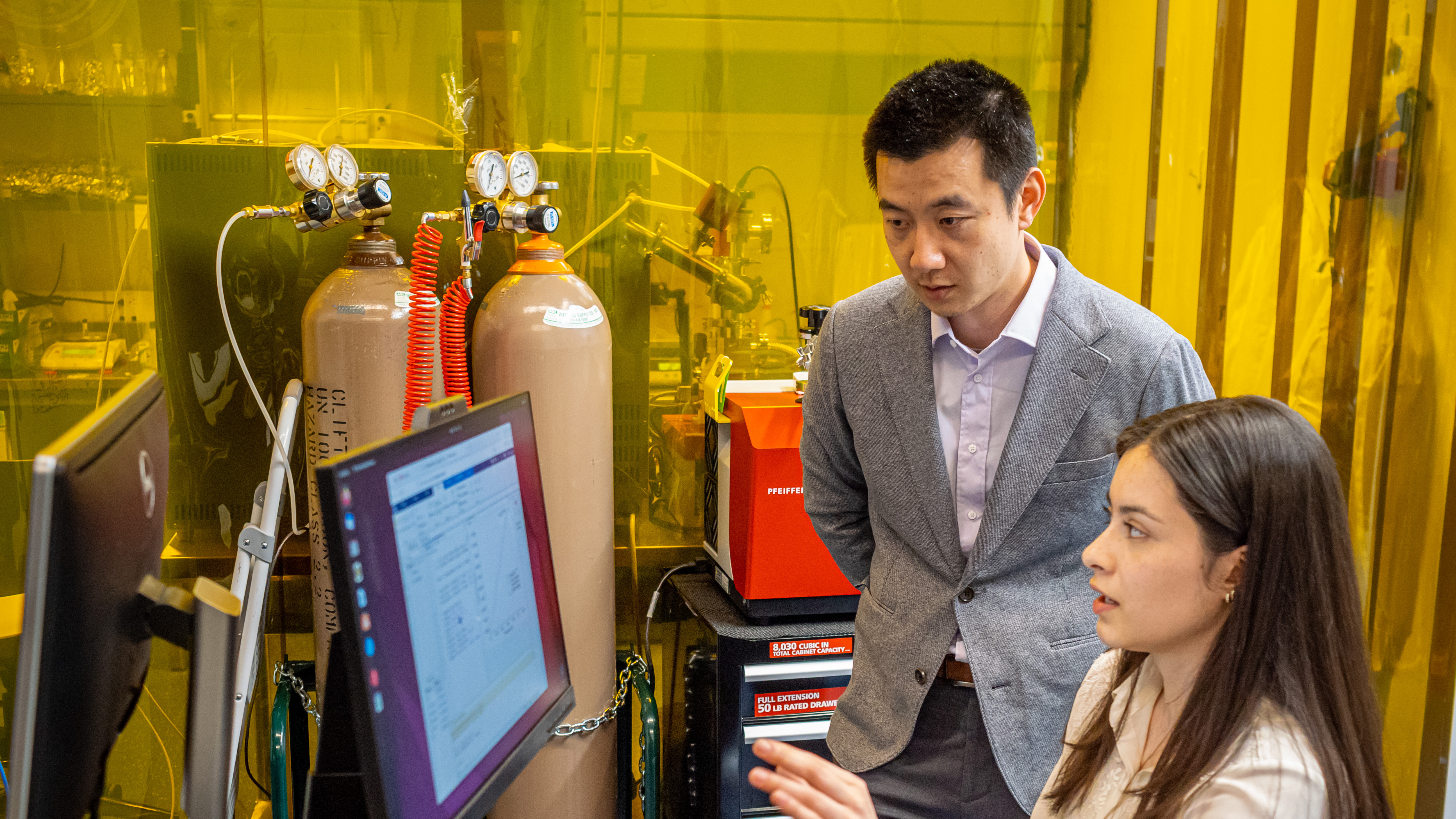 A seated woman gestures at a computer monitor as Professor Kent Leung looks on
