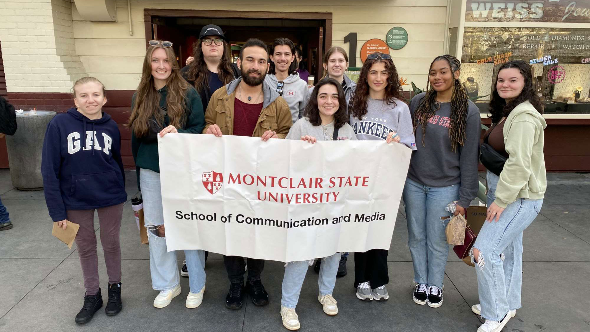Semester in LA students pose for a photo in front of the Los Angeles Farmers Market, holding a banner that says "Montclair State University School of Communication and Media."