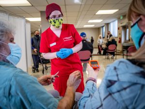 nurse wearing PPE talking with two seniors who are seated in a waiting area
