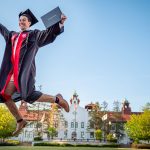 graduate in cap-and-gown leaping through the air with diploma in hand