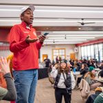 A man stands on a table and addresses a group of students.
