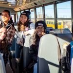 Six female students smile at the camera while sitting on bus seats.
