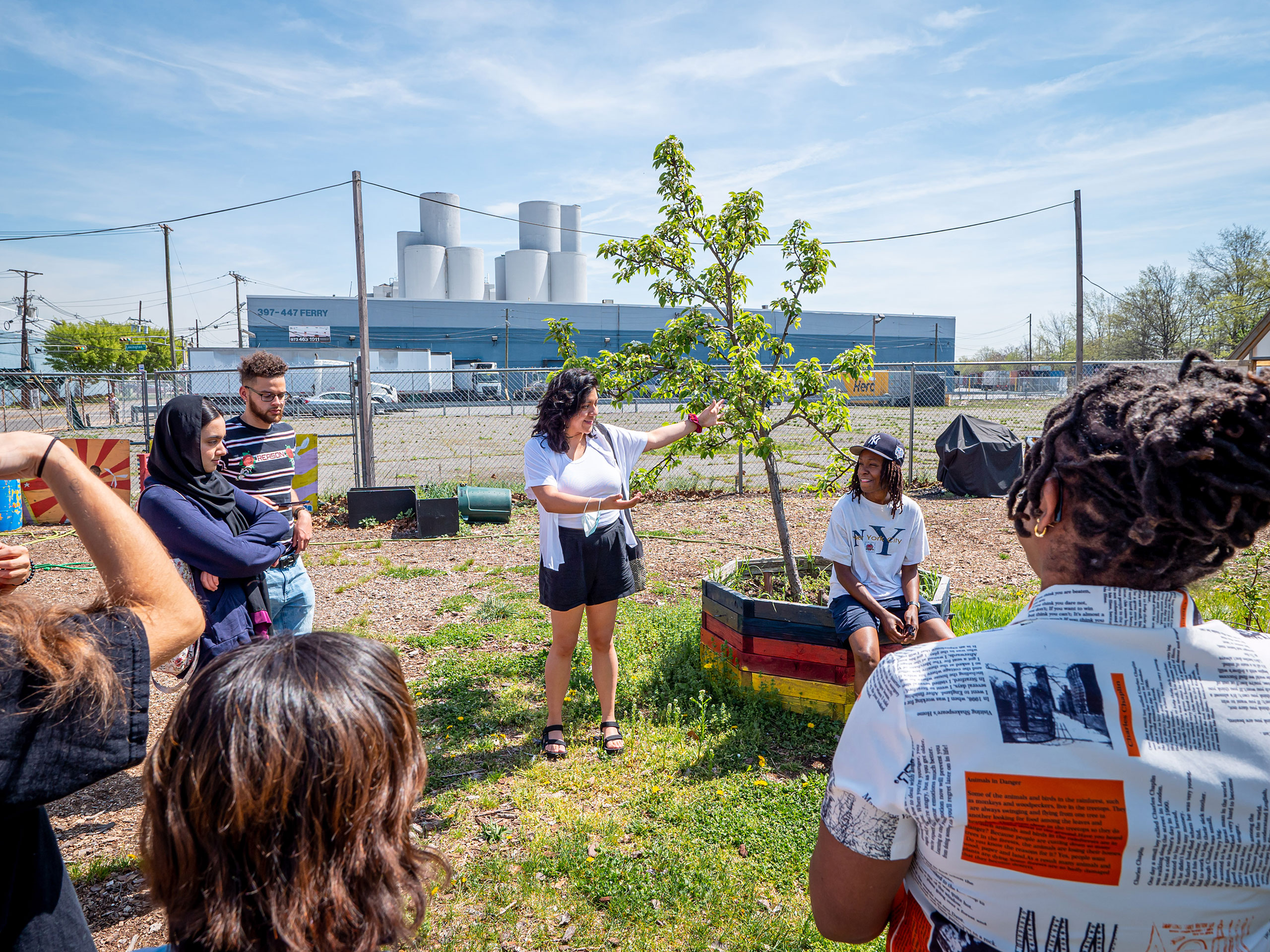 A factory visible in the background as a tour guide next to a tree speaks to a group of people at a community garden.