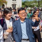 Korean faculty member and two Korean students pose for selfie on the Student Center patio while a crowd of event attendees moves about behind them.